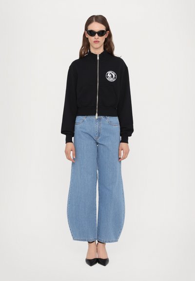 Black zip-up sweatshirt with a logo patch, paired with wide-leg light blue jeans and black pointed-toe heels. Overall casual outfit.