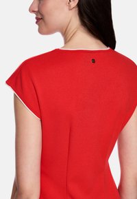 Rotes Kurzarmshirt in tailliertem Design mit weißem Besatz entlang des Halsausschnitts und kleinem schwarzem Logo auf der Rückseite. Glatte Textur.