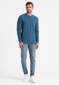 Mann steht in einem türkisfarbenen Langarm-Henley-Shirt, hellblauen Jeans und weißen Sneakers vor einem weißen Hintergrund.