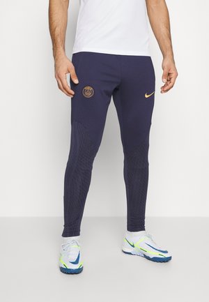 Mann trägt marineblaue Paris Saint-Germain Nike Trainingshose und weiße Sportschuhe mit blauen und neongelben Akzenten.