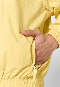 Lys gul jakke lavet af blødt stof, med elastisk talje, lommer og justerbare snore til tilpasning.