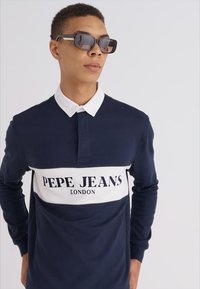 Polo cu mânecă lungă, de culoare bleumarin, cu o bandă de accent albă pe care scrie, cu litere îngroșate, "PEPE JEANS LONDON". Modelul poartă ochelari de soare închiși. Textura fină.