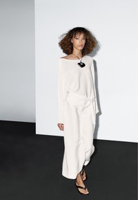 Donna che cammina indossando un top bianco a spalle scoperte e una gonna lunga, con una collana pendente nera e sandali neri, su uno sfondo bianco e nero.