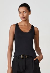Donna che indossa un top nero senza maniche con bordo in pizzo, pantaloni neri e una cintura nera con fibbia dorata, in piedi con una mano in tasca.
