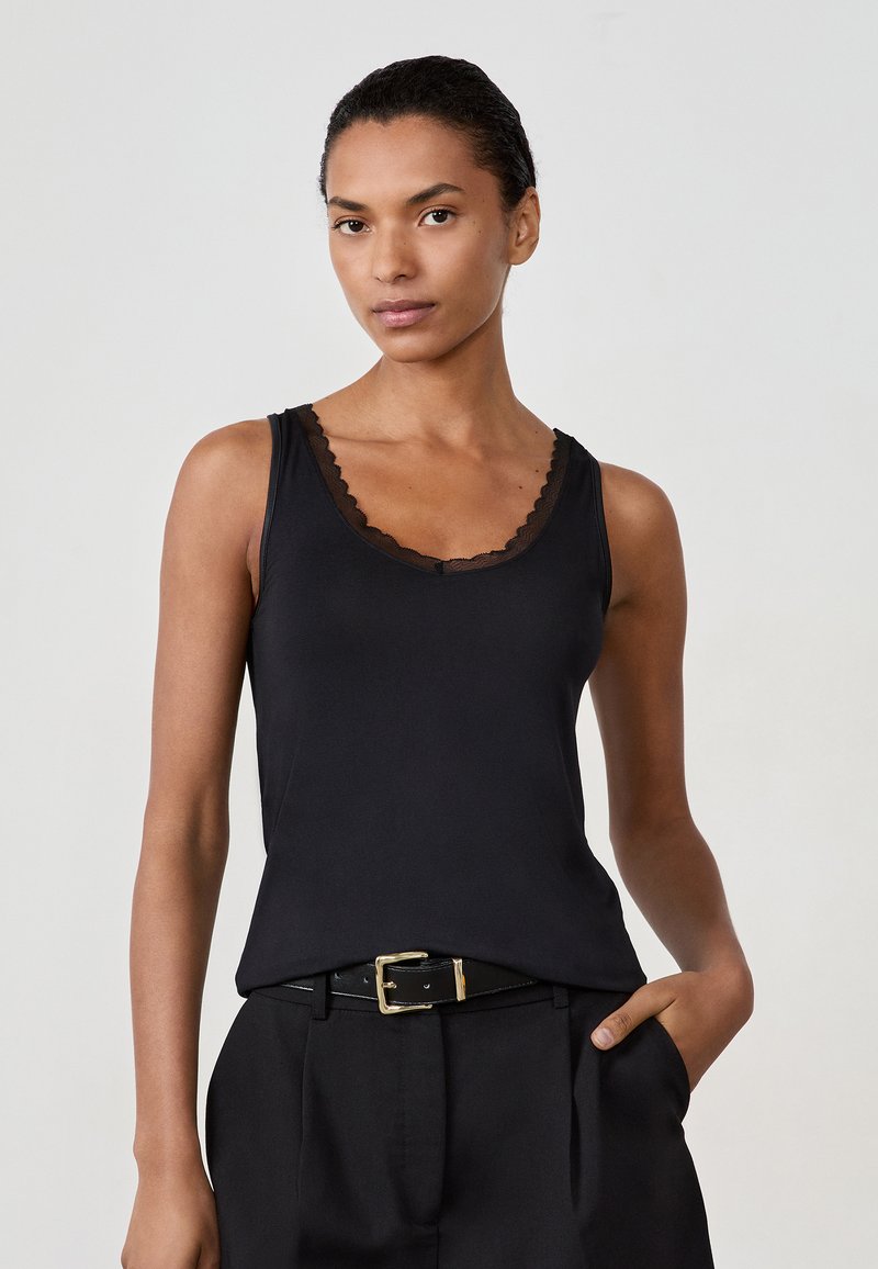 Donna che indossa un top nero senza maniche con bordo in pizzo, pantaloni neri e una cintura nera con fibbia dorata, in piedi con una mano in tasca.