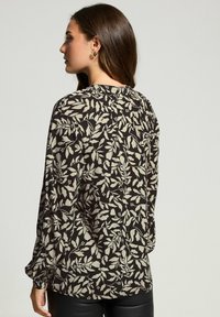 Frau mit langen braunen Haaren trägt eine schwarze Bluse mit beigem Blumenmuster, von hinten vor einem schlichten hellen Hintergrund gezeigt.