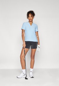 Polo à manches courtes bleu clair avec col en V, motif texturé, associé à un short noir et des chaussures de golf blanches, tenant un club de golf.