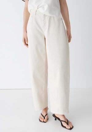 Persona che indossa pantaloni crema a vita alta e gamba larga abbinati a sandali neri con tacco e una maglietta bianca infilata, in piedi su un pavimento bianco.