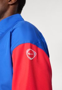 Közeli kép egy személyről, aki kék és piros dzsekit visel, a piros ujja lebegő Nike logóval fehér hímzéssel.