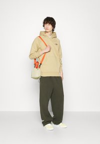 Felpa beige con tasca frontale e logo, pantaloni in pile verdi, sneakers bianche con dettagli gialli e una piccola borsa leggera con tracolla arancione.