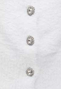 Λευκό χνουδωτό ένδυμα με τρία διακοσμητικά κουμπιά με καθαρά κρύσταλλα ραδιονίων τοποθετημένα σε ασημί υλικό. Απαλή υφή και απλός σχεδιασμός.