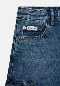 Calvin Klein plave traperice s malim prednjim džepom, vidljivim šavovima i crvenom oznakom. Tkanina pokazuje blago izblijedeli izgled.