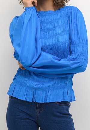 Person iført en lyseblå smockbluse med lange ærmer og flæsekanter samt mørkeblå jeans, arm bøjet med hånd nær ansigtet.