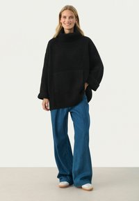 Zwarte, oversized gebreide trui met een hoge nek en een voorzak, gecombineerd met blauwe denim broeken met wijde pijpen en witte sneakers.