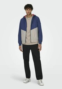 Marineblauwe en grijze windbreaker met een rits aan de voorkant, contrasterende kleurvlakken, elastische manchetten en capuchon. Draagt over een beige shirt en zwarte broek.