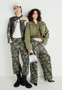 Δύο άτομα στέκονται δίπλα-δίπλα. Το άτομο στα αριστερά φοράει ένα μαύρο δερμάτινο μπουφάν, λευκό πουκάμισο και καμουφλαρισμένα παντελόνια. Το άτομο στα δεξιά φοράει ένα πράσινο κοντό μπουφάν και καμουφλαρισμένα παντελόνια.