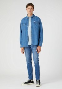 Ljust blå denimskjorta med två bröstfickor, lagd över en ljusblå skjorta med knappar, ihop med mörkblå jeans och svarta sneakers.
