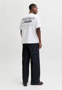 Λευκό βαμβακερό T-shirt με μαύρο κείμενο στην πλάτη, συνδυασμένο με φαρδιά σκούρα τζιν και καφέ παπούτσια. Απλός, σύγχρονος σχεδιασμός.