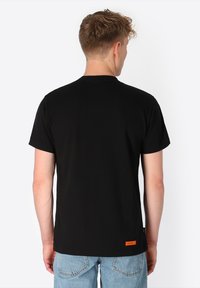 Junger Mann mit kurzen braunen Haaren, der ein schlichtes schwarzes T-Shirt mit kurzen Ärmeln und hellblauen Jeans trägt und von der Kamera abgewandt ist.
