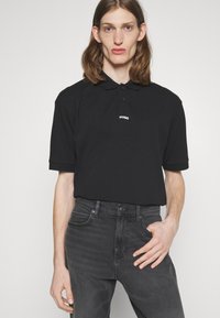 Tricou polo negru cu o deschidere cu trei butoane și logo-ul alb "HUGO", asortat cu blugi gri închis, care au cinci buzunare și o croială ajustată.