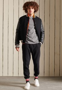Zwart varsity jack met leren mouwen, oranje en witte accenten, grijs T-shirt, zwarte trainingsbroek en witte sneakers met rode en zwarte strepen.