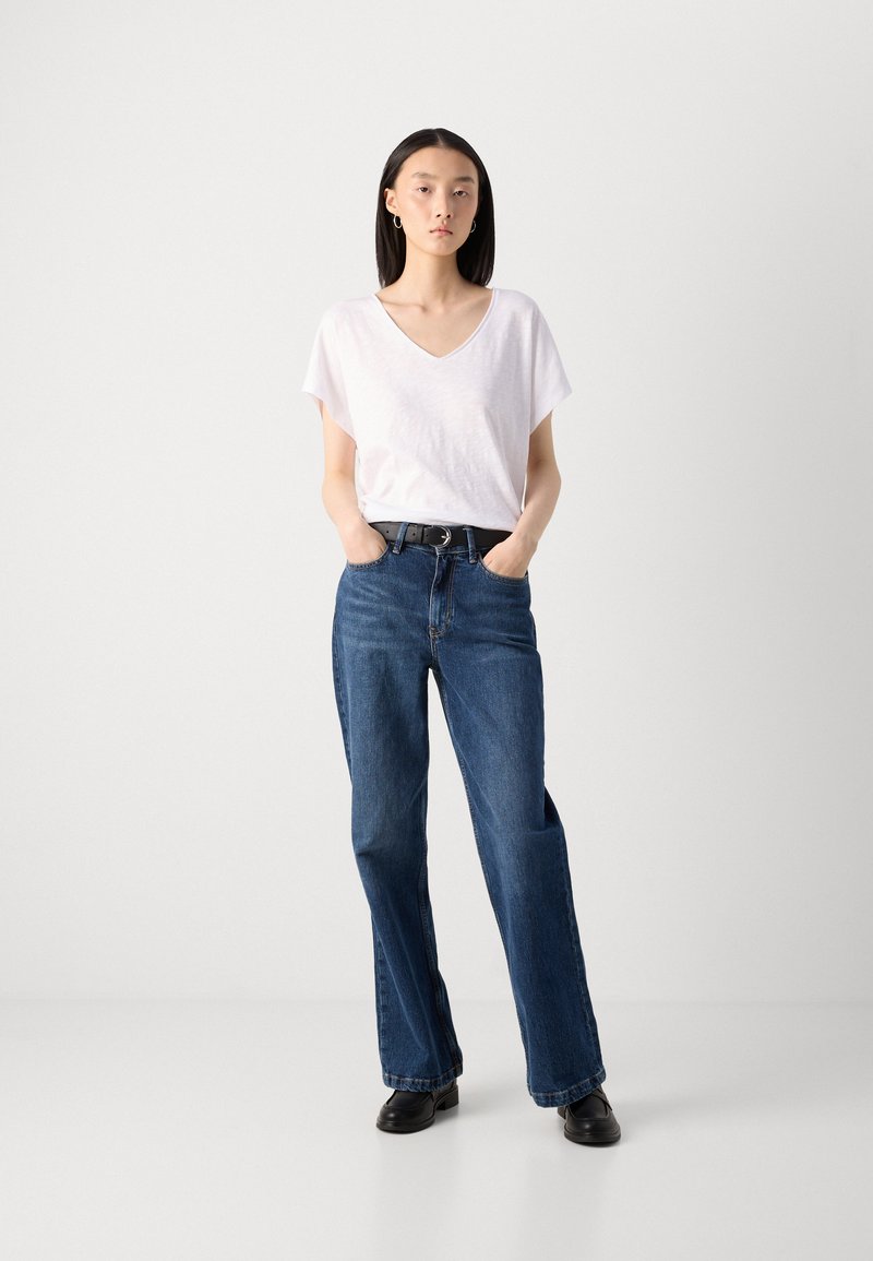 Biała koszulka z dekoltem w serek, luźny krój, w połączeniu z ciemnoniebieskimi dżinsami prostymi. Model nosi czarny pasek i czarne buty. Prosty design, bez wzorów.