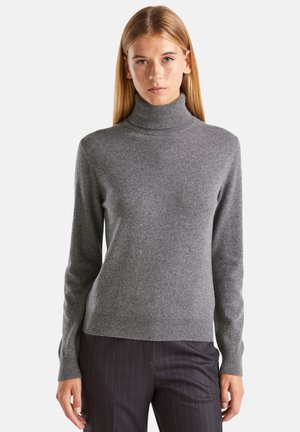 Jeune femme portant un pull col roulé gris et un pantalon à rayures foncées, se tenant devant un fond blanc uni.