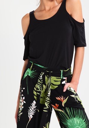 Femme portant un top noir à épaules dénudées et un pantalon noir avec un imprimé feuilles et fleurs tropicales vertes et oranges, main dans la poche.