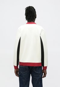 Sudadera blanca con cuello y dobladillo acanalados en rojo, con paneles laterales negros y mangas largas. Textura suave, ajuste casual y detalles mínimos.