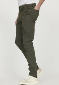 Pantalons vert khaki à coupe slim, dotés de poches, d'un tissu texturé et de détails de couture subtils, portés avec des baskets blanches et beiges.
