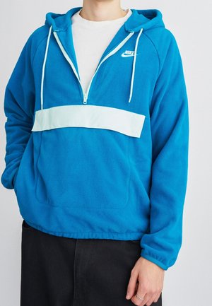 Personne portant un sweat à capuche en polaire bleu Nike avec une bande blanche sur la poitrine et une fermeture éclair, assorti à un pantalon noir et un sous-vêtement blanc.