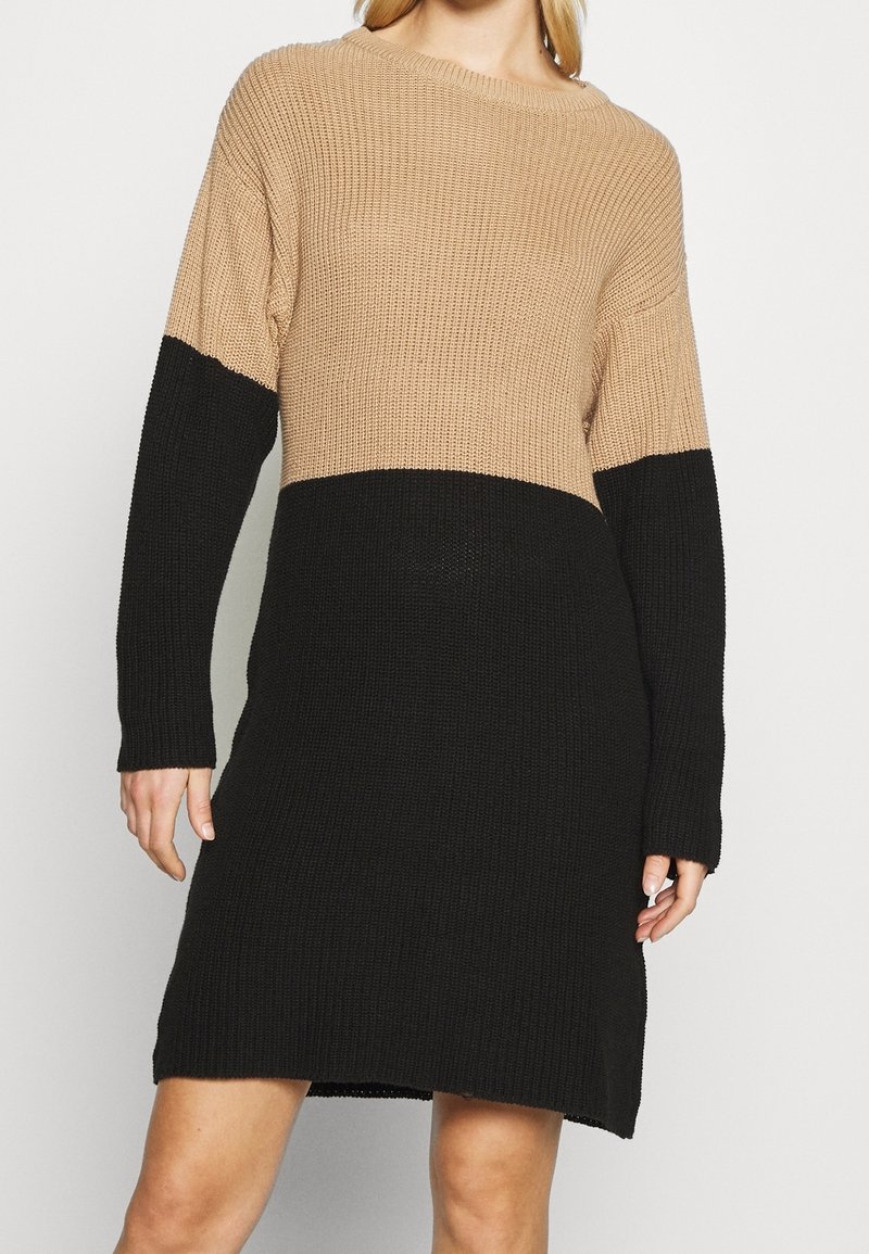 Stickad klänning i beige och svart. Har en ribbad struktur, långa ärmar och en avslappnad passform, med en färgblockerad design vid midjan.