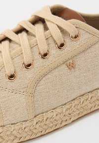 Bež platnena tenisica s tkanim jutenim potplatom, koja ima zlatne okove, ravne vezice i mali logotip s strane.
