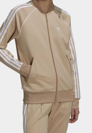 Person iført beige Adidas træningsjakke og bukser med hvide striber og logo, hånd i jakkelommen, foran ensfarvet baggrund.