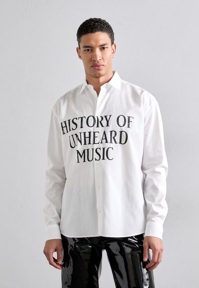 Chemise blanche à boutons avec un texte noir indiquant "HISTOIRE DE LA MUSIQUE INAUDITE." Tissu lisse, manches longues et col classique.