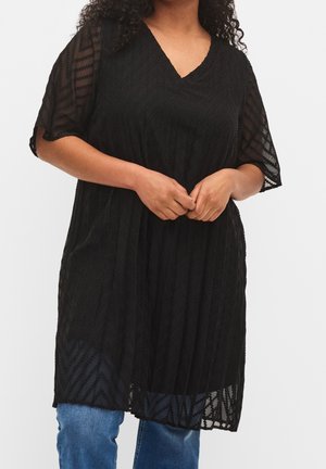 Tunique noire en tissu transparent avec un motif zigzag texturé, dotée d'un col en V et de manches courtes. Portée sur un jean bleu.