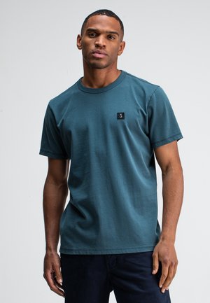 Mann, der ein einfaches türkisfarbenes T-Shirt mit kurzen Ärmeln und einem kleinen schwarzen Logo-Patch auf der linken Brust trägt, steht vor einem weißen Hintergrund.