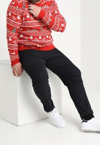 Pull de Noël rouge avec motifs blancs, associé à un pantalon noir et des baskets blanches. Main posée sur la poitrine, assis sur un cube blanc.