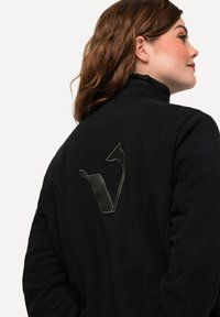 Černá fleecová bunda s vysokým límcem a výrazným grafickým designem v šedé barvě na zadní straně, který zobrazuje abstraktní tvar.