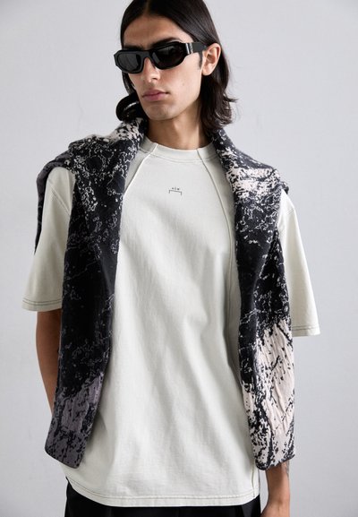 Rövid ujjú, világosszürke ing, fekete-fehér sállal, amelyen nyomtatott minta található, kiegészítve szögletes fekete napszemüveggel.