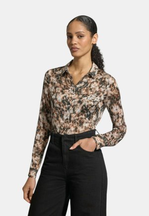 Donna con capelli ricci raccolti in una coda indossa una camicetta a maniche lunghe con bottoni decorata con un motivo astratto marrone e nero e pantaloni neri a vita alta.