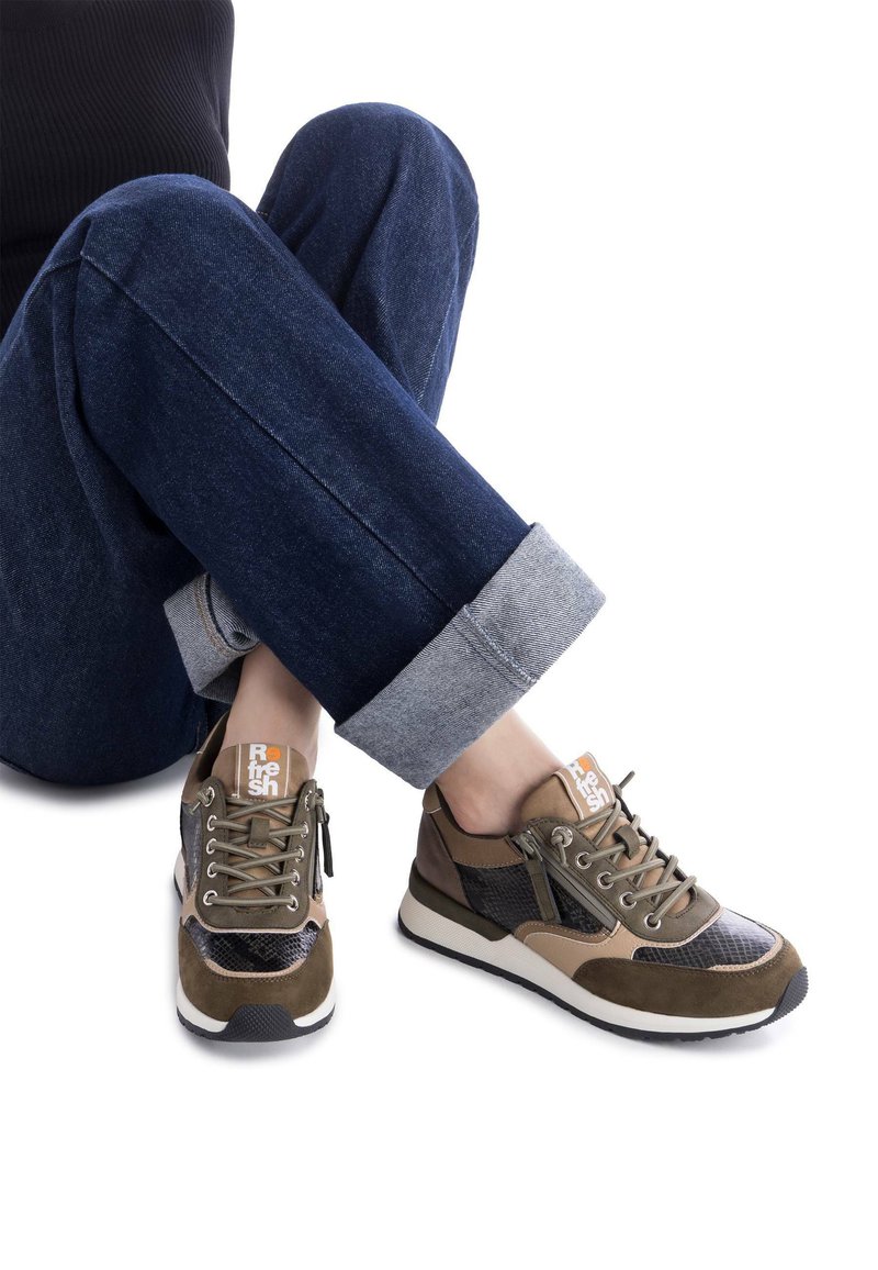 Zapatillas marrones y verde oliva con acentos de malla y ante. Suela blanca, cordones y una etiqueta de logo en la lengüeta. Jeans oscuros de tiro recto.