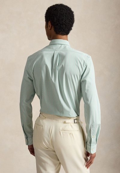 Homme portant une chemise à manches longues rayée vert clair et blanc rentrée dans un pantalon beige, vu de dos sur un fond neutre.