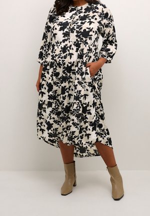 Vrouw draagt een zwart-witte midi-jurk met bloemenprint en driekwartmouwen, gecombineerd met beige enkellaarzen, staand met één hand in haar zak.