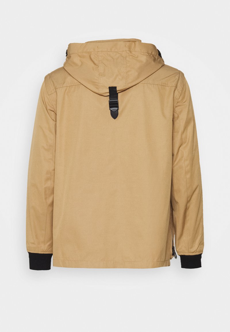 Veste à capuche beige avec une texture lisse, présentant un accent noir aux poignets et un accessoire en forme de boucle à l'arrière pour l'accrocher.