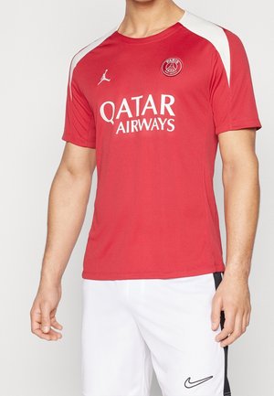 Czerwona koszulka piłkarska Paris Saint-Germain z logo Qatar Airways oraz białe spodenki z czarnym logo Nike, noszone przez stojącą osobę.