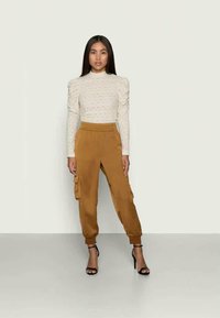 Κρέμ πλεκτό τοπ με φουσκωτά μανίκια, συνδυασμένο με χαλαρά παντελόνια cargo σε μουσταρδί απόχρωση με πλαϊνές τσέπες. Το μοντέλο φορά μαύρα τακούνια.