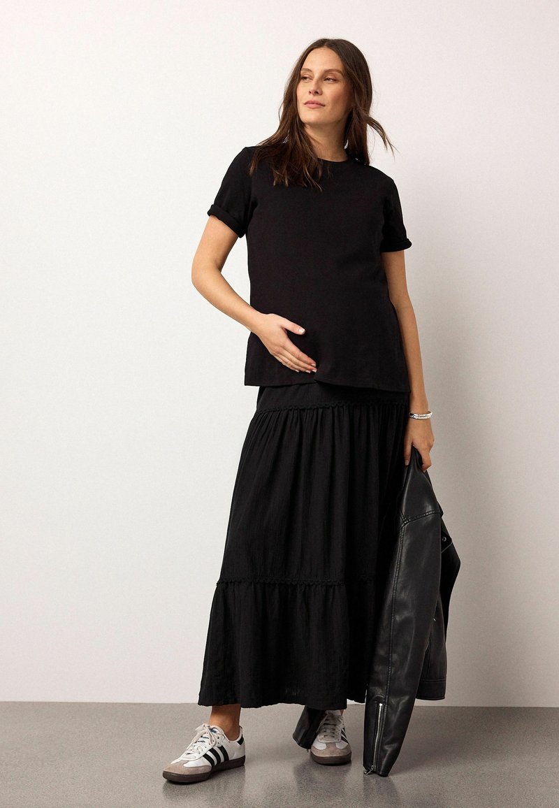 Femme enceinte portant un haut noir à manches courtes et une jupe longue, tenant une veste en cuir noire dans une main, chaussée de baskets blanches.
