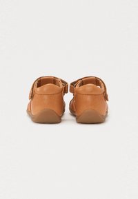 Chaussures en cuir tan avec une forme arrondie, une sangle arrière et une surface texturée. La semelle est d'un brun clair uni, conçue pour le confort.