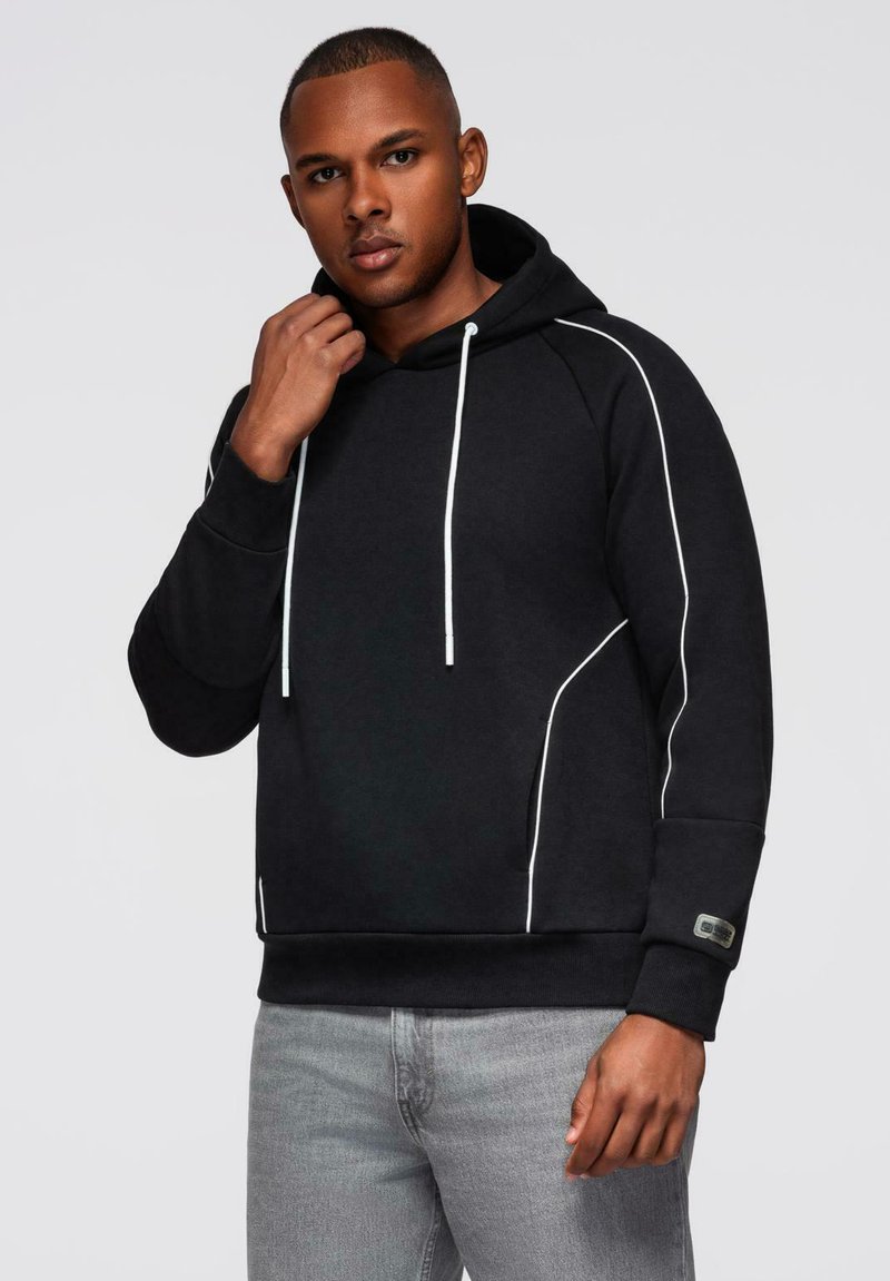 Mann in einem schwarzen Kapuzenpullover mit weißen Abnähten und Kordelzügen, der mit einer Hand an seinem Hals posiert und graue Jeans trägt.